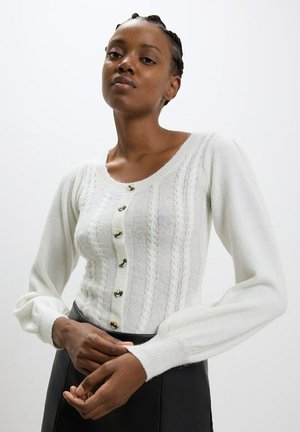 Maglione bianco lavorato a maglia con un motivo testurizzato, scollatura rotonda e bottoni argentati sul davanti. Maniche lunghe a sbuffo con polsini a costine.