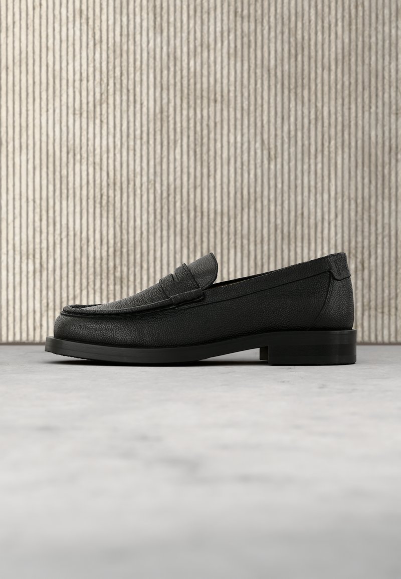 Mocassin homme en cuir noir avec bout arrondi et petit talon, présenté sur un fond à lignes verticales texturées sur un sol lisse.