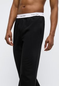 Μαύρο βαμβακερό παντελόνι πιτζάμα με λευκή ελαστική ζώνη μέσης που φέρει το λογότυπο "Calvin Klein". Λείο ύφασμα, ίσια γραμμή με πλαϊνές τσέπες.