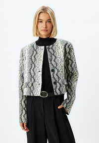 Veste cropped avec un motif serpent gris et blanc. Équipée de cinq boutons, d'un col rond et de deux poches avant.