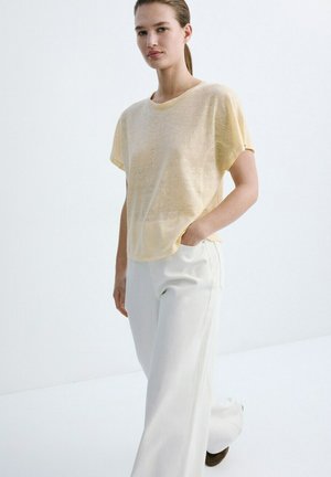 Ženska z lasmi, povezanimi nazaj, oblečena v ohlapno bledo rumeno majico s kratkimi rokavi in širokimi belimi hlačami, stoji z eno roko v žepu.