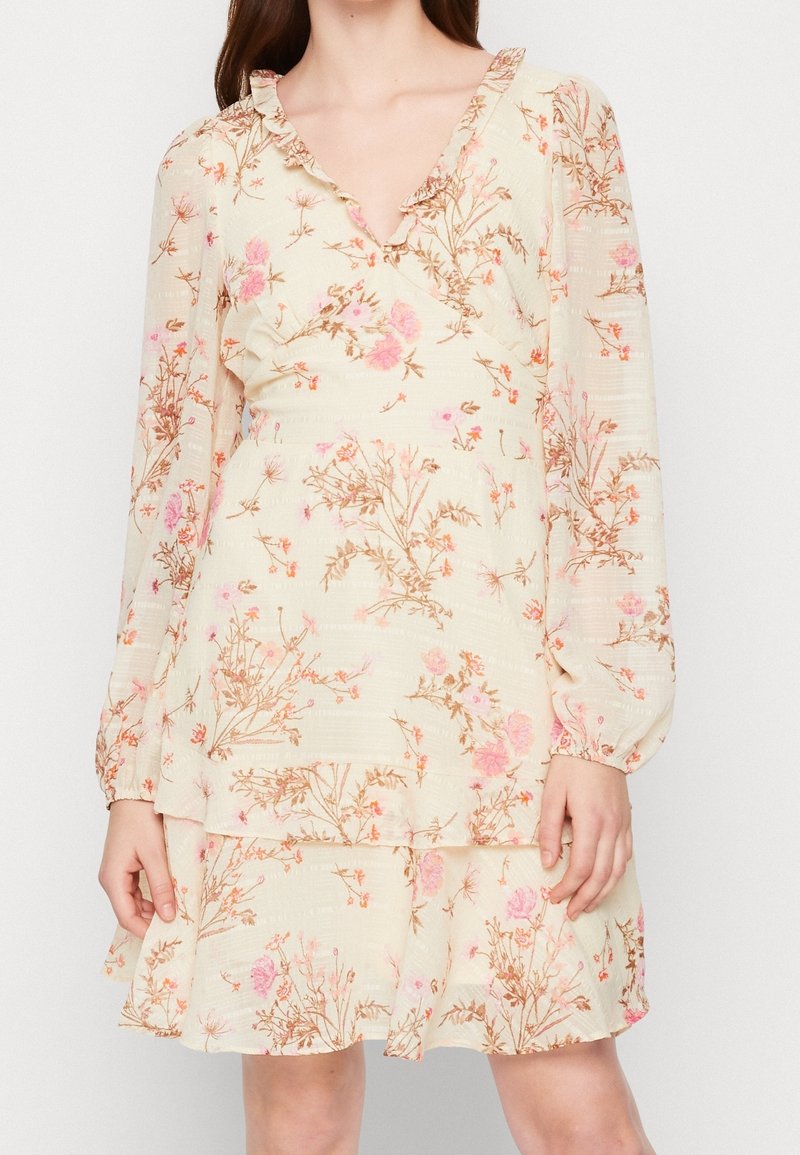 Femme portant une robe florale de couleur crème avec des fleurs roses et orange, des manches longues bouffantes, un col en V et un ourlet volanté.