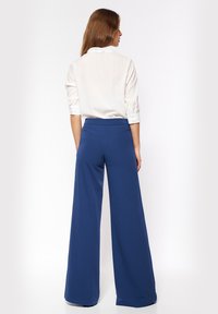 Kobieta z długimi brązowymi włosami, ubrana w białą bluzkę i niebieskie spodnie o wysokim stanie i szerokich nogawkach, stojąca tyłem na białym tle.