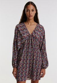 Kurzes Blumenkleid in Schwarz mit bunten Blumen. V-Ausschnitt, lange Ärmel und plissierte Textur. Der Stoff wirkt leicht und fließend.