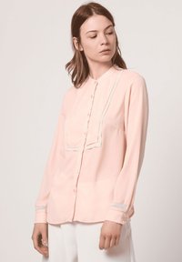Blusa de color rosa claro con cuello redondeado, cierre frontal de botones y acentos decorativos de encaje a lo largo de la tapeta y en los puños. Tejido suave.