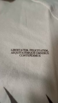 Tessuto bianco con testo in rilievo di colore marrone scuro: "LIBERTATEM, FELICITATEM, AEQUITATEMQUE OMNIBUS CONTENDIMUS." Texture liscia, design minimale.