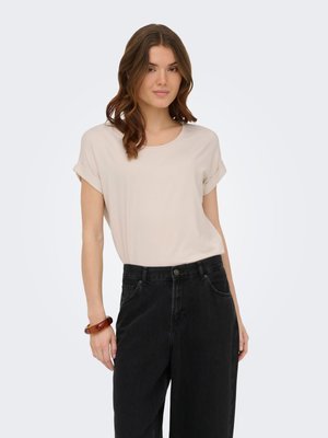 Jeune femme aux cheveux bruns ondulés portant un top beige à manches courtes, un pantalon noir taille haute et un bracelet marron, debout devant un fond blanc.