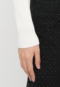 Λευκό, ριγωτό, μακρυμάνικο τοπ, δίπλα σε μια μαύρη φούστα από υφή με μικρές ανυψωμένες κουκίδες, επιδεικνύει αντίθετα υλικά και χρώματα.