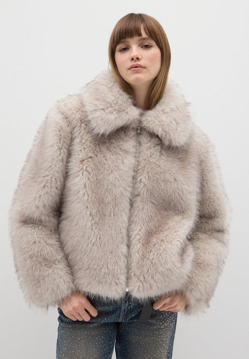 Nepbonten jas in lichtbeige met een brede kraag, ritssluiting aan de voorkant en een zachte textuur. Gecombineerd met donkere jeans met kleine zilveren stippen.