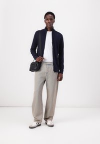 Navy zip-up jacket over a white t-shirt, paired with light gray wide-leg jeans and black bag. White sneakers with black accents complete the look.