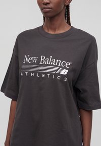 Άτομο που φοράει ένα σκουρόγκρι φαρδύ T-shirt με τη φράση «New Balance Athletics» και λογότυπο σε λευκό στο μπροστινό μέρος, στέκεται μπροστά σε απλό φόντο.