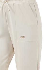 Pantaloni jogger beige realizzati in un tessuto morbido e liscio. Presentano un elastico in vita con cordino, tasche laterali e un piccolo logo dorato "EAZ" sulla tasca.