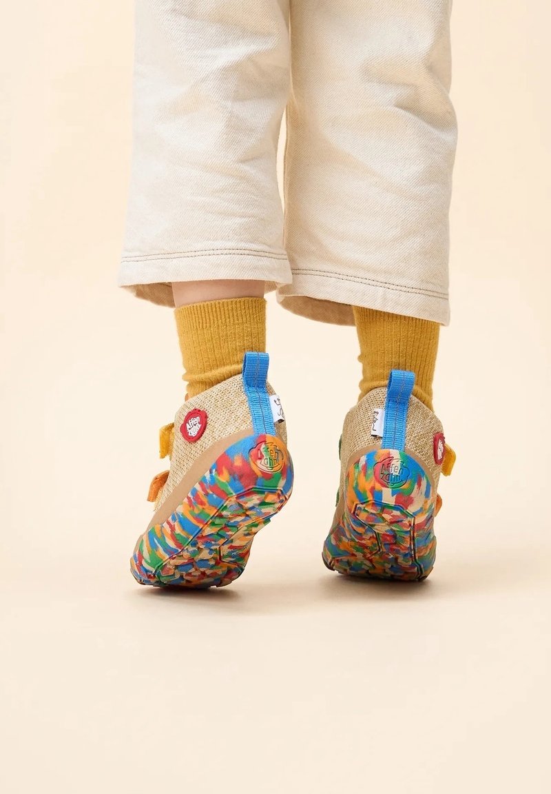 Enfant portant un pantalon beige, des chaussettes moutarde et des baskets colorées avec des semelles multicolores et des languettes bleues, debout sur un fond clair.