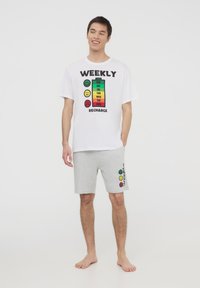 T-shirt bianco in cotone con una grafica di una batteria colorata e la scritta "RICARICA SETTIMANALE". Pantaloni corti in jersey grigi con un motivo di faccine sorridenti sul lato.