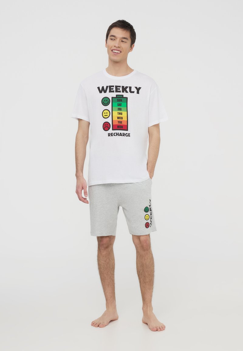 T-shirt bianco in cotone con una grafica di una batteria colorata e la scritta "RICARICA SETTIMANALE". Pantaloni corti in jersey grigi con un motivo di faccine sorridenti sul lato.