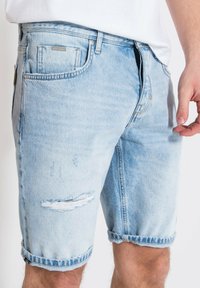 Pantalones cortos de denim azul claro con un ajuste holgado, dobladillos enrollados y detalles desgastados con parches deshilachados en la parte delantera.