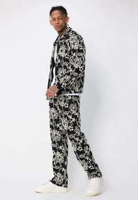 Homme debout portant une veste noire et un pantalon avec des motifs floraux blancs, ainsi que des baskets blanches, sur fond uni.