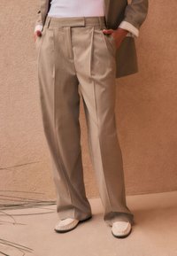 Pantaloni beige chiaro su misura con pieghe frontali, tasche laterali e una texture liscia. Abbinati a scarpe bianche slip-on.