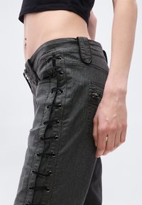 Σκούρα γκρι τζιν με δέσιμο στα πλάγια, που διαθέτουν μεταλλικά κρικάκια και λεία υφή. Κοντινή λήψη στο πως εφαρμόζουν και στη θέση του χεριού.