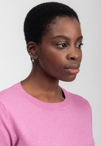 Maglione rosa a maglia con scollo rotondo. Indossato da una persona con capelli neri corti, che sfoggia orecchini d'argento con perle e accenti decorativi.