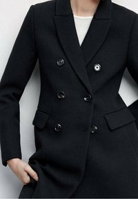 Vrouw in een nauwsluitende zwarte dubbelgeknoopte blazer met zes knopen, twee voorste klepzakken en een witte top eronder.