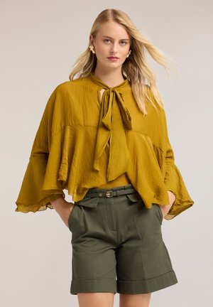 Blonde vrouw draagt een losse mosterdkleurige blouse met strik en olijfgroene short met riem, handen in de zakken, tegen een neutrale achtergrond.