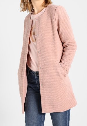 Femme portant un long cardigan texturé rose clair sur un t-shirt graphique rose et un jean bleu, main dans la poche, sur un fond blanc.