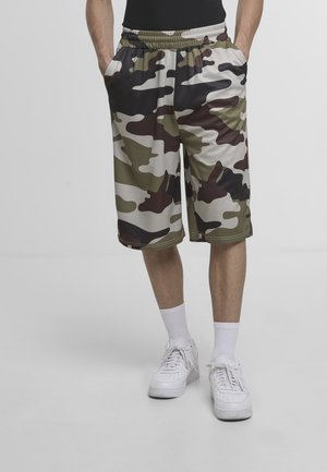 Mężczyzna ubrany w zielone, czarne i brązowe krótkie spodenki moro z kieszeniami do kolan, białe trampki i białe skarpety do kostek, stoi z rękami w kieszeniach.