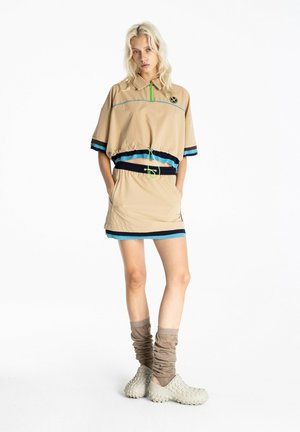 Mujer con un top corto beige con rayas azules y negras, falda a juego, calcetines taupe caídos y zapatos beige sin cordones con textura, de pie con las manos en los bolsillos.