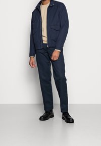 Chaqueta de marina con cierre delantero, cuello a presión y múltiples bolsillos, combinada con un suéter beige y pantalones marinos a juego, además de zapatos negros.