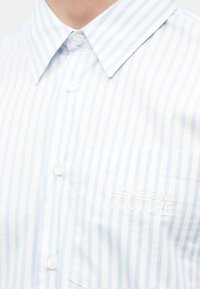 Galléros, világoskék és fehér csíkos, gombos ing mellkasi zsebbel, amelyen enyhén fehér hímzésben olvasható a "Versace Jeans Couture" felirat.