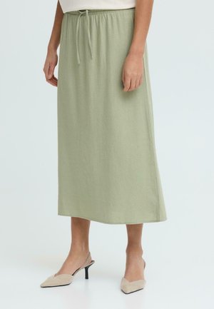 Kvinde iført en salviegrøn midi-nederdel med elastisk snøre i taljen og beige spidse slingback-hæle, stående på en hvid gulv.