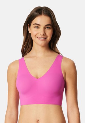 Uśmiechnięta kobieta z długimi brązowymi włosami, ubrana w jaskraworóżowy, bezrękawnikowy sportowy biustonosz z dekoltem w kształcie litery V na jasnym, jednolitym tle.