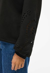 Felpa nera con maniche lunghe caratterizzata da design floreali intagliati sui polsini. Tessuto morbido, vestibilità rilassata, abbinata a jeans blu.