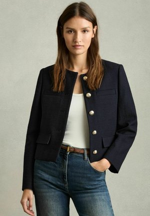 Marineblauer Cropped-Blazer aus strukturiertem Stoff, mit goldenen Knöpfen, zwei Vordertaschen und rundem Ausschnitt, kombiniert mit einem weißen Oberteil.
