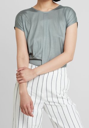 Kvinde iført en lysegrå top med korte ærmer og højtaljede hvide bukser med tynde lodrette sorte striber, armene krydset foran taljen.