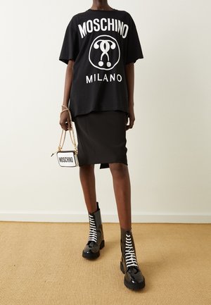 Personne portant un t-shirt noir surdimensionné Moschino Milano, une jupe noire longueur genou, des bottes à lacets en cuir verni noir, tenant un sac blanc Moschino avec une chaîne dorée.