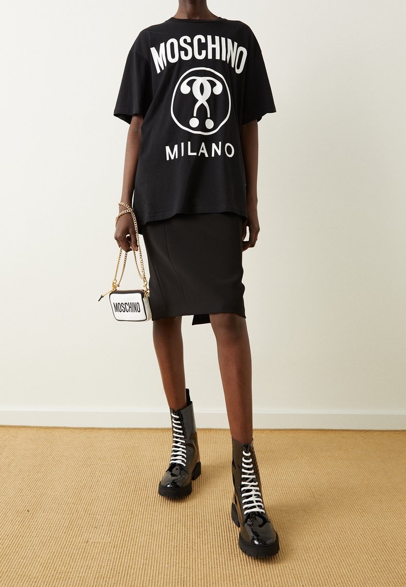 Persona que lleva una camiseta negra de gran tamaño de Moschino Milano, falda negra hasta la rodilla, botas negras de charol con cordones, sosteniendo un bolso blanco de Moschino con cadena dorada.