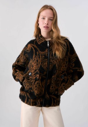 Junge Frau mit langen, welligen Haaren, die eine schwarz-braun gemusterte, flauschige Reißverschlussjacke mit großen Knöpfen und helle Hose vor schlichtem Hintergrund trägt.