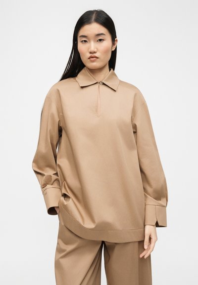 Femme asiatique aux longs cheveux noirs portant une chemise ample à col beige à manches longues et un pantalon large assorti, debout devant un fond blanc.