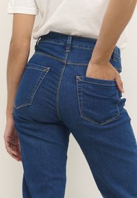 Des jeans en denim bleu taille haute présentent des poches arrière avec une texture subtile et une couture jaune contrastante. Le tissu semble durable et lisse.