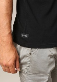 Gros plan sur la main d'une personne reposant à côté d'un pantalon gris clair et d'un t-shirt noir avec une petite étiquette "Deeluxe Original" près de l'ourlet.