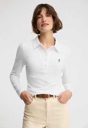 Frau trägt ein weißes langärmeliges Poloshirt mit Knöpfen, beige Hose und einen braunen Gürtel, steht mit den Händen in den Taschen.