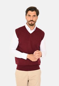 Maglione senza maniche bordeaux con scollatura a V, realizzato in materiale morbido, indossato sopra una camicia bianca a maniche lunghe, abbinato a pantaloni color khaki.