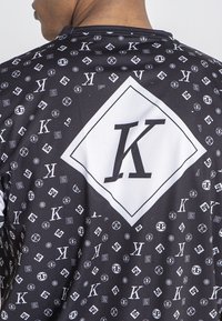 Camisa de tela negra con un patrón de logotipos repetidos. Gran forma de diamante blanca con una "K" en negrita en la parte posterior; textura suave.