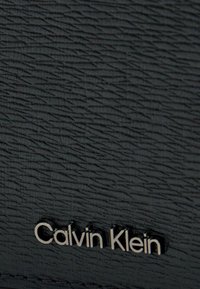 Cuero negro con textura que presenta un patrón en relieve y un logotipo metálico plateado de "Calvin Klein" en la parte inferior.