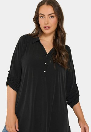 Femme aux longs cheveux bruns portant une chemise noire boutonnée avec les manches retroussées sur un fond clair uni.