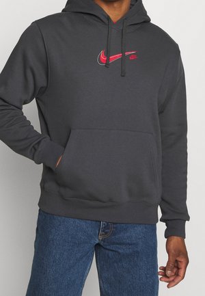 Mężczyzna w ciemnoszarej bluzie z kapturem z czerwonym logo Nike i niebieskich dżinsach, ręka w przedniej kieszeni, stoi na tle jednolitego tła.