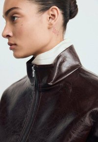 Brown leather jacket with high collar and zip closure, featuring a textured finish. White turtleneck visible underneath.