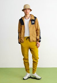Nike-Jacke in Braun und Schwarz mit Logo, kombiniert mit gelben, taillierten Sweatpants. Das Outfit umfasst einen beigefarbenen Bucket Hat und Sneakers mit strukturierter Oberfläche.